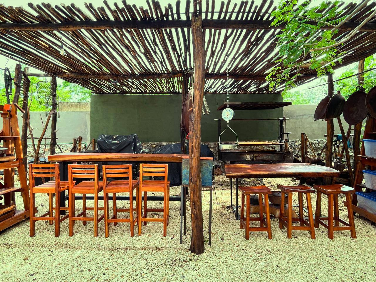 Un patio exterior con techo de madera, mesas y sillas de madera, y una parrilla.