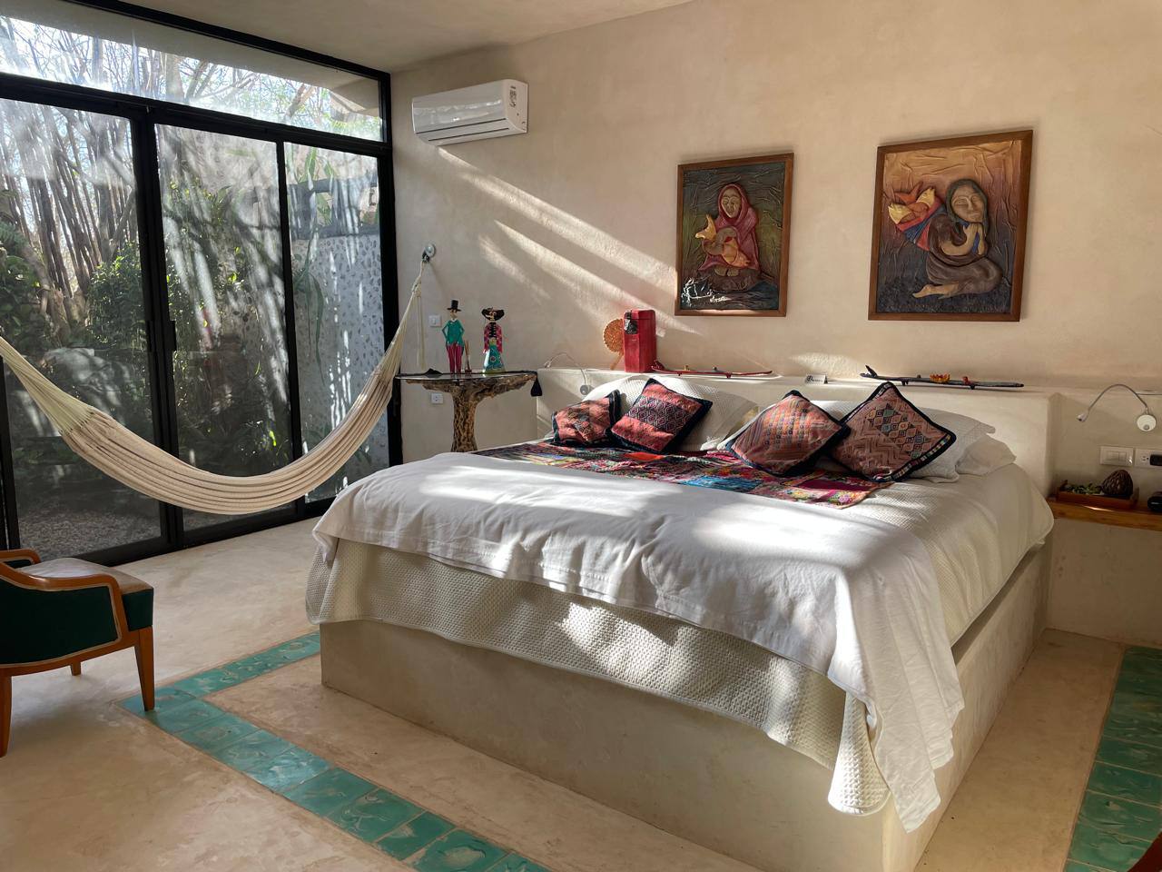 Recámara con cama grande, hamaca, ventanal al jardín y decoración artesanal en la pared.