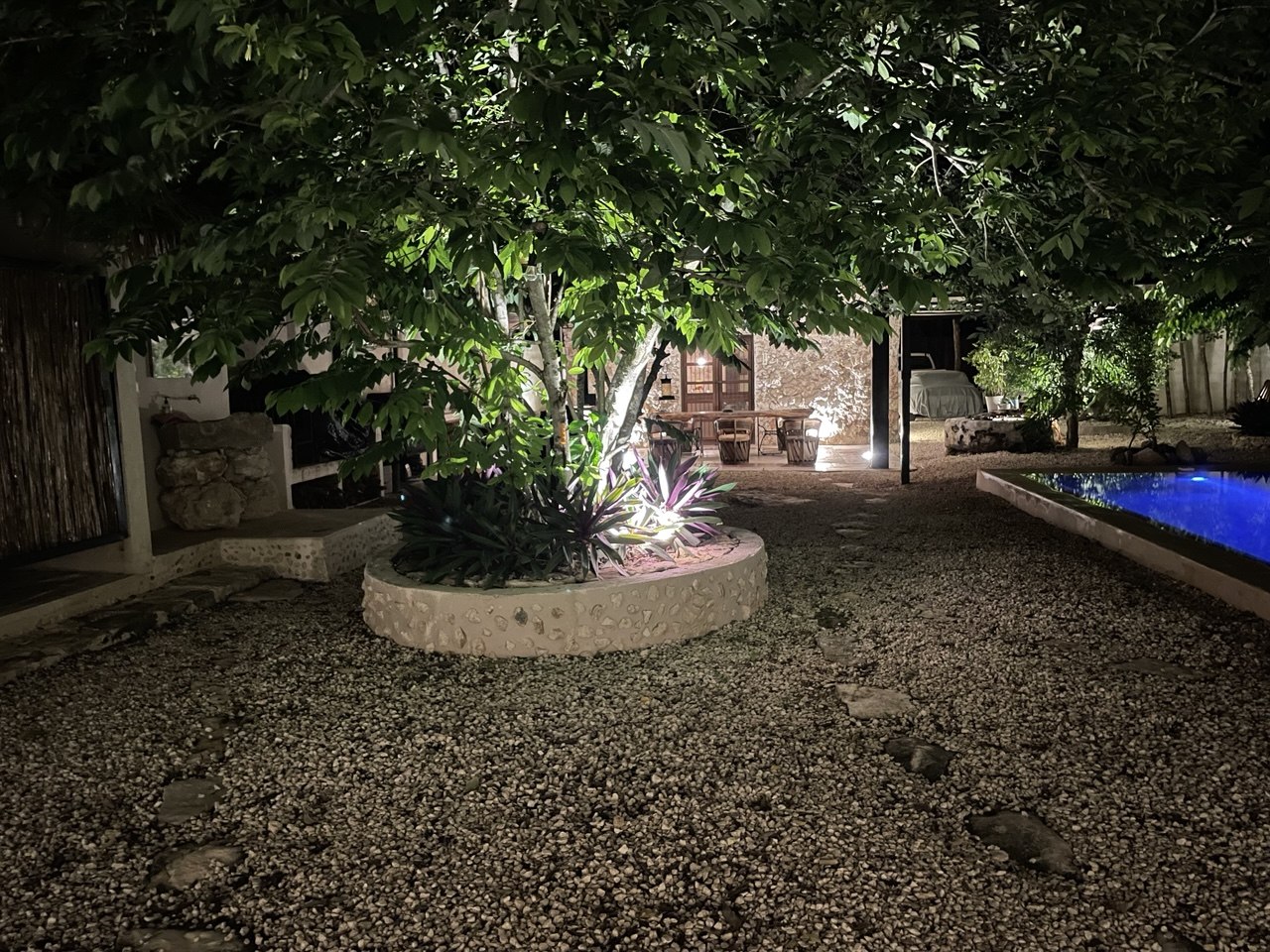 Patio exterior con árboles iluminados, camino de grava, piscina y área de comedor.