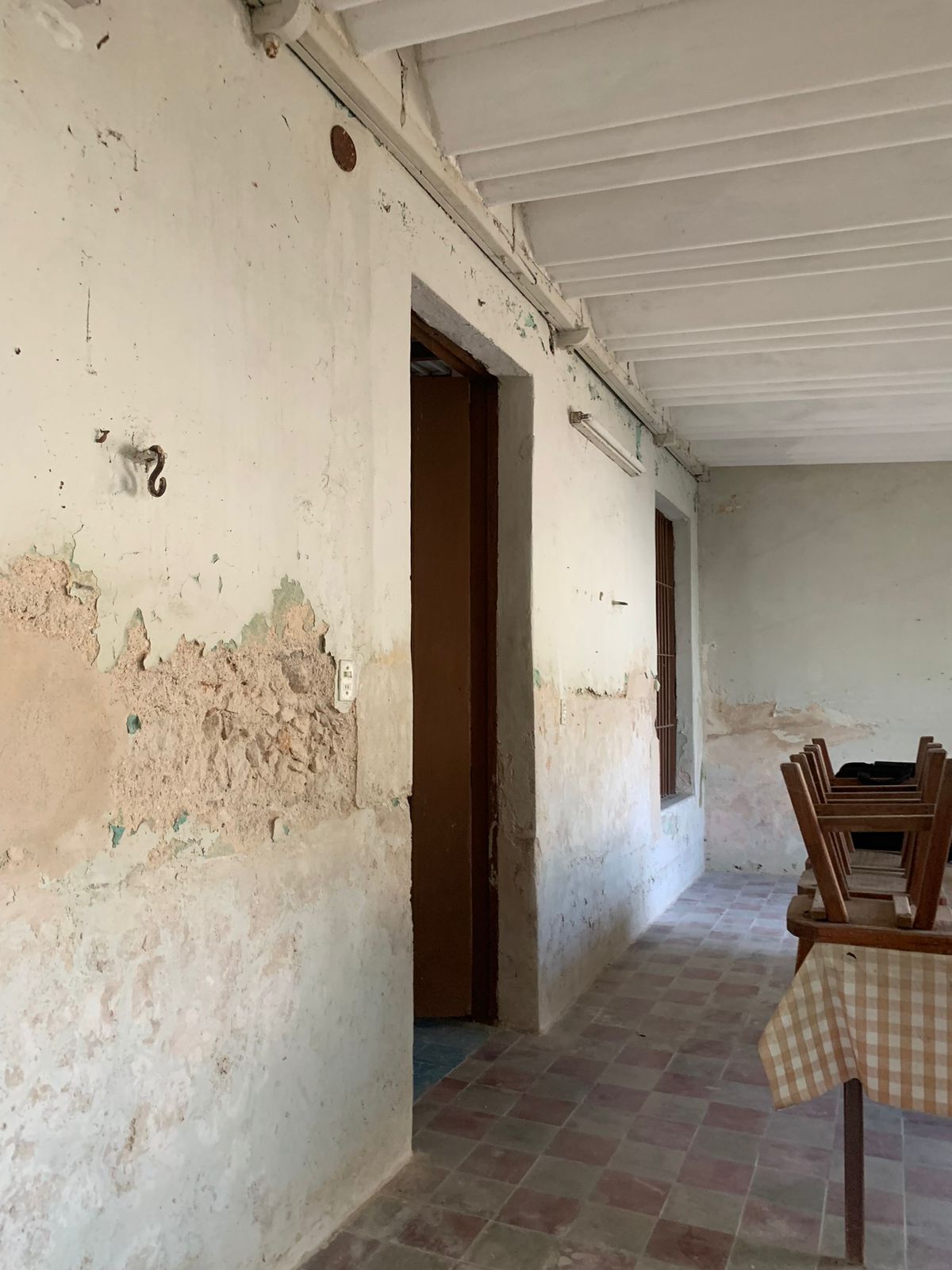 Habitación con paredes deterioradas, techo de vigas, piso de baldosas y sillas apiladas.