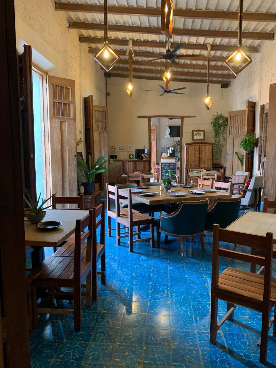 Comedor rústico con mesas y sillas de madera, piso azul y lámparas colgantes.