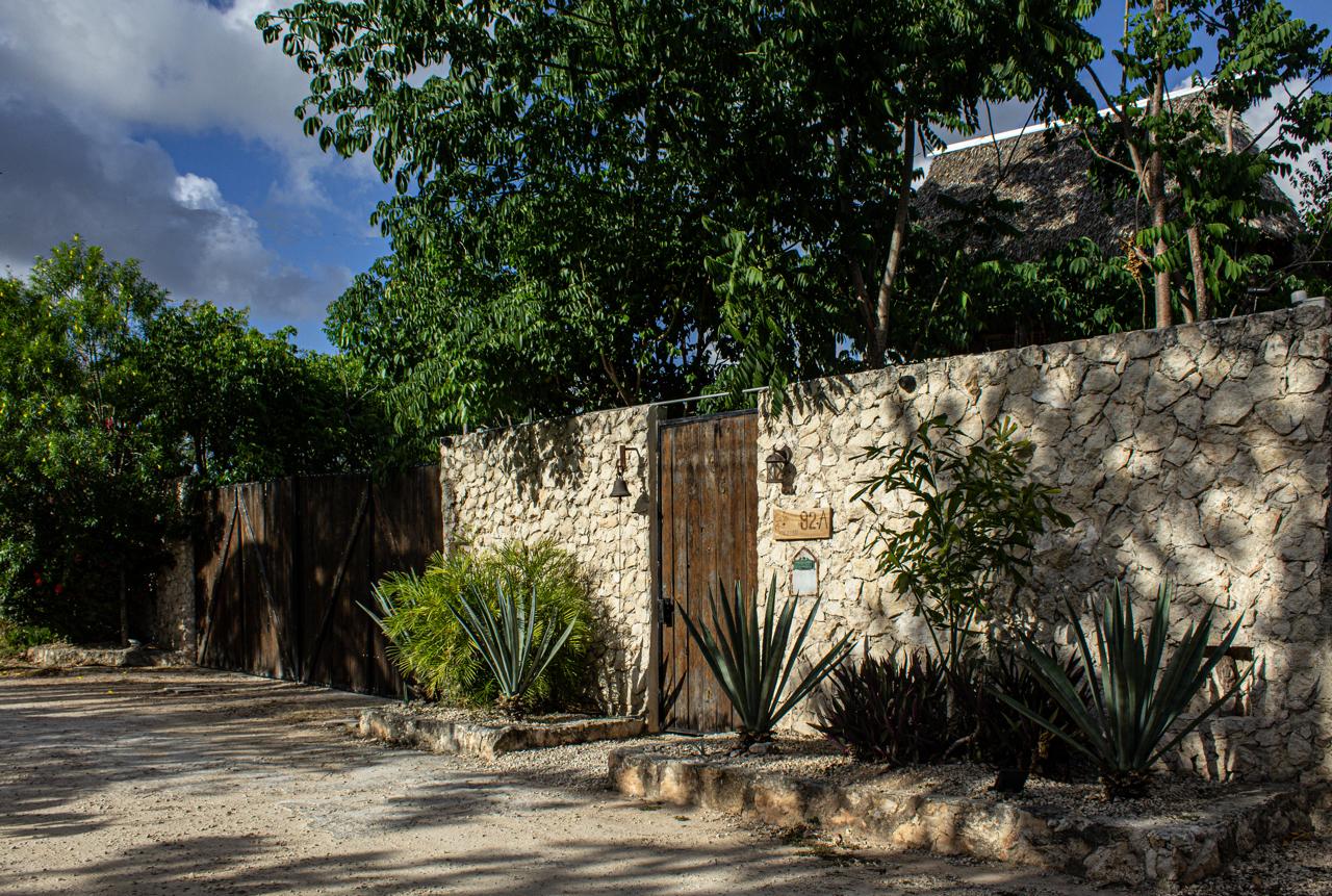 Muro de piedra con puerta de madera, plantas y árboles bajo un cielo nublado.