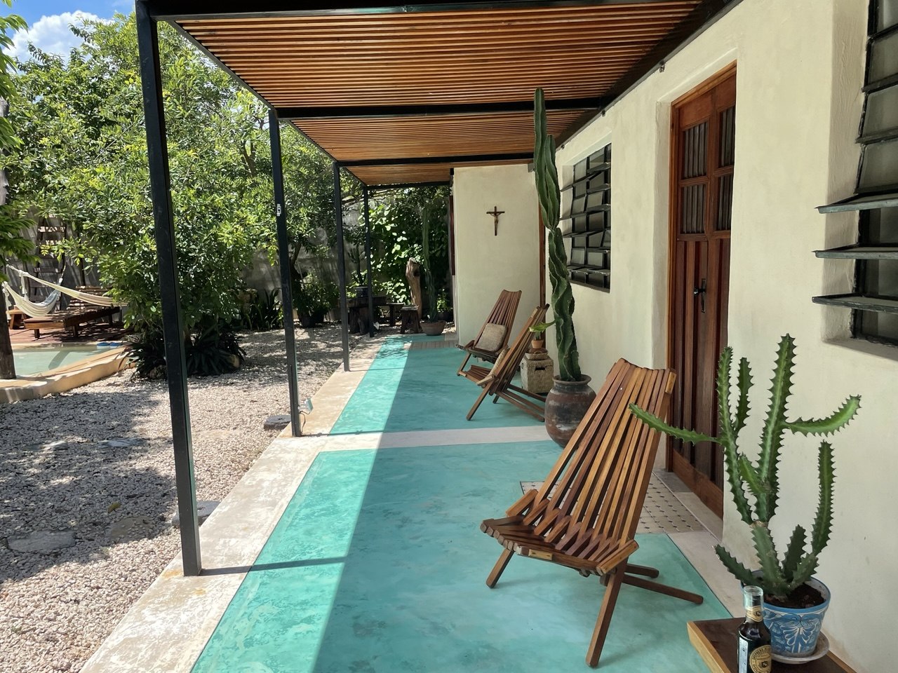 Patio exterior con techo de pérgola, sillas de madera, plantas y hamaca al fondo.