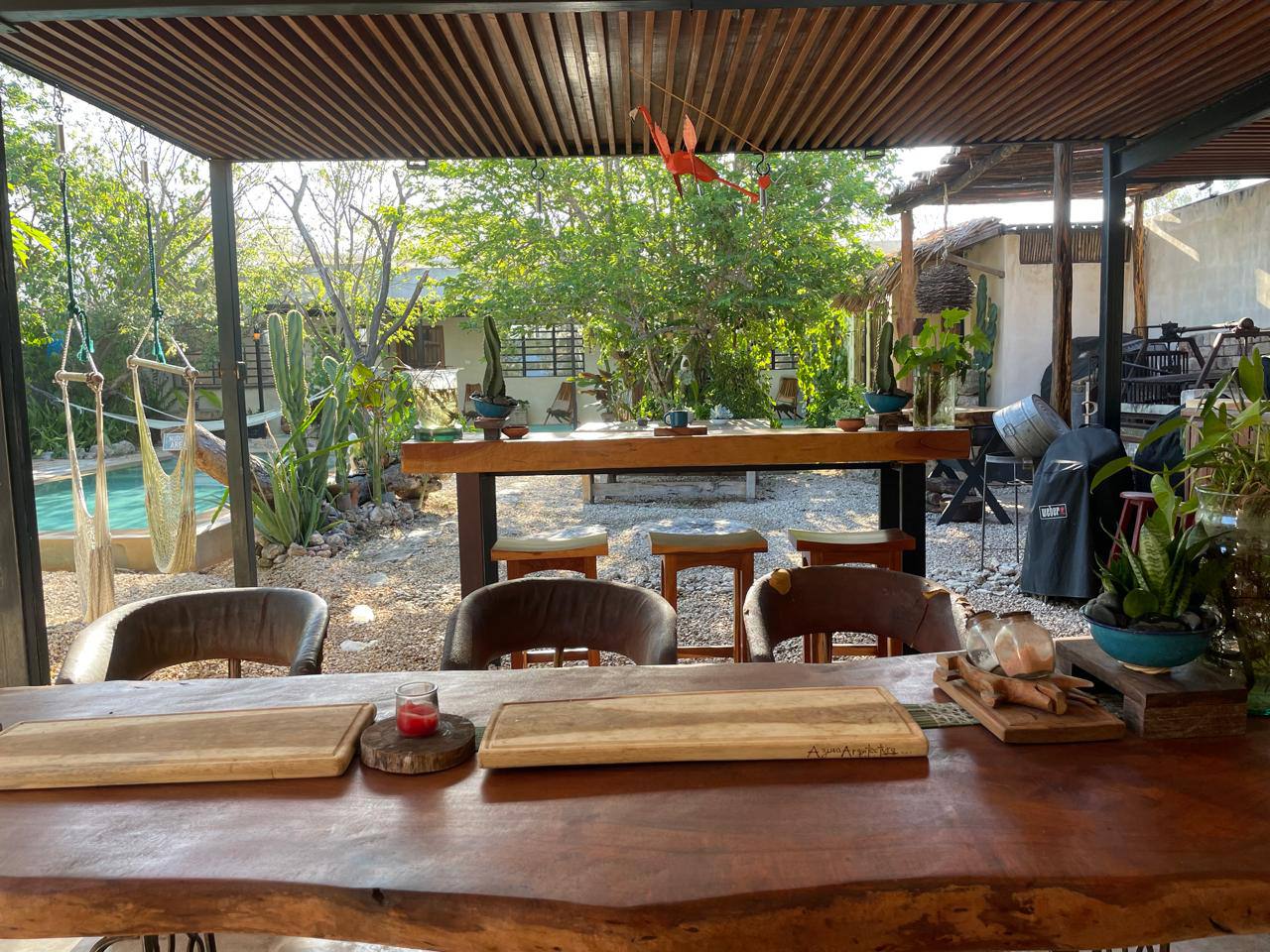 Patio exterior con mesa de madera, sillas, barra, plantas y alberca al fondo.