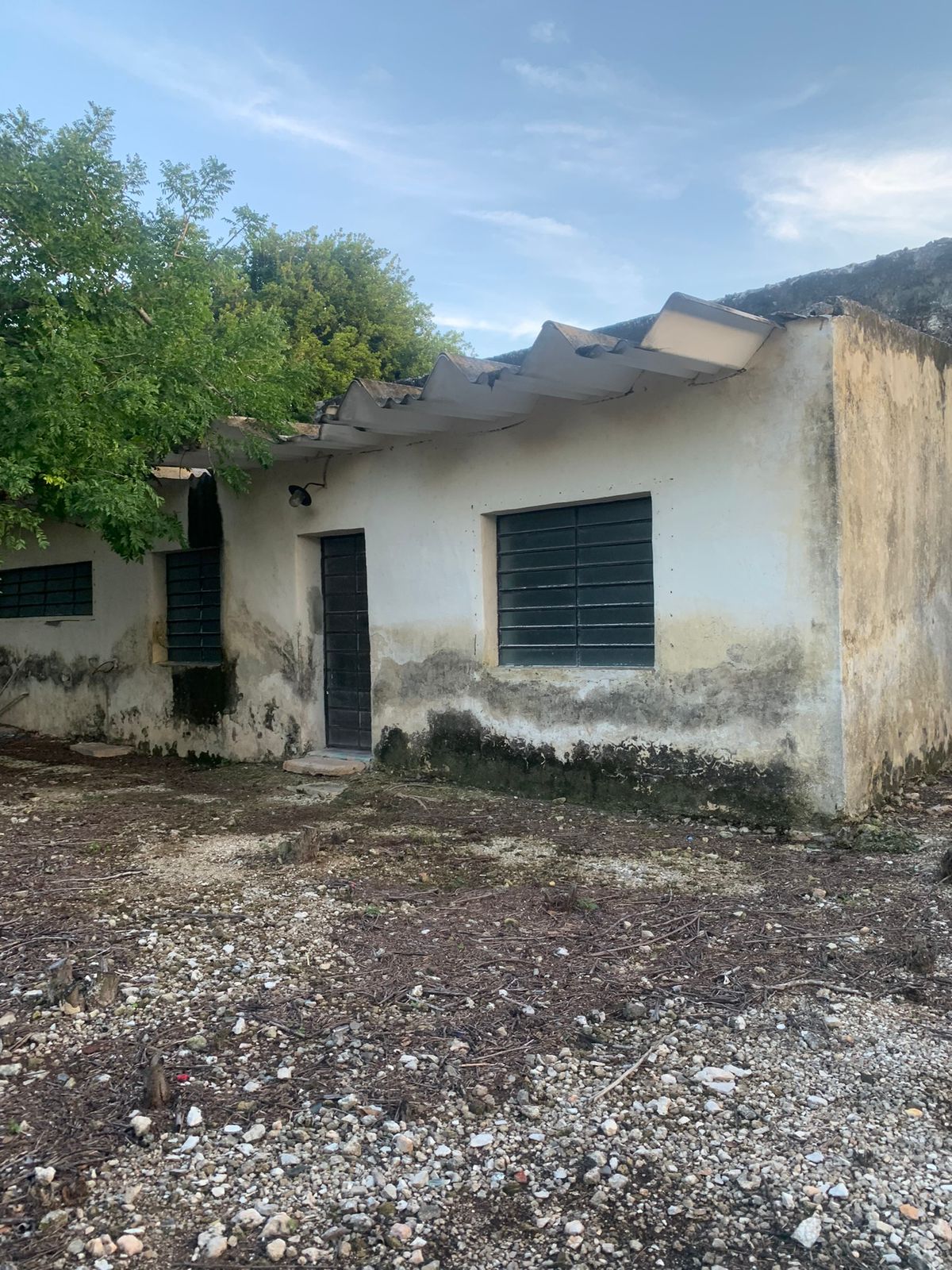 Exterior de una casa antigua con techo de lámina, paredes sucias y terreno pedregoso.