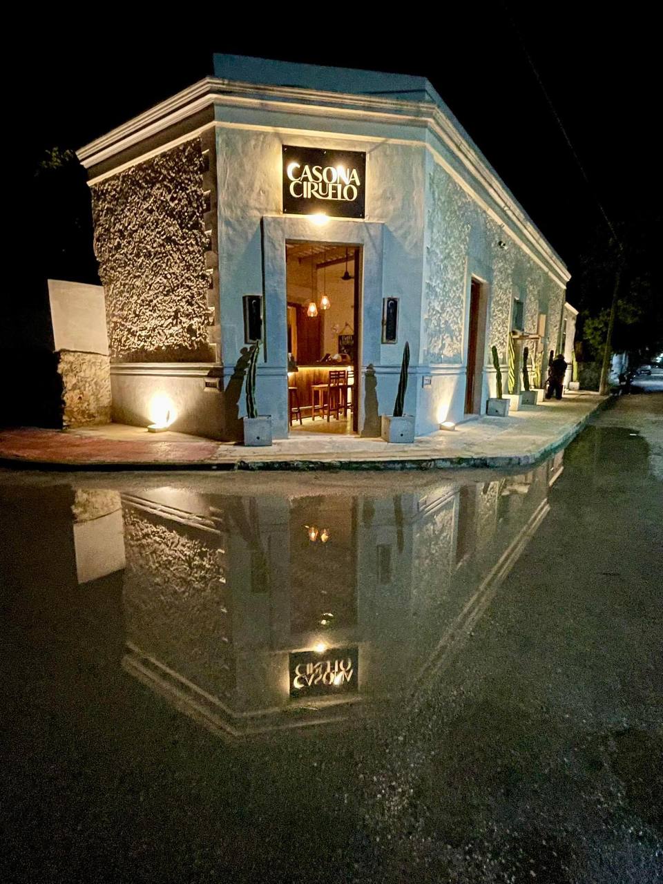 Fachada de "Casona Ciruelo" iluminada de noche, con su reflejo en un charco de agua.