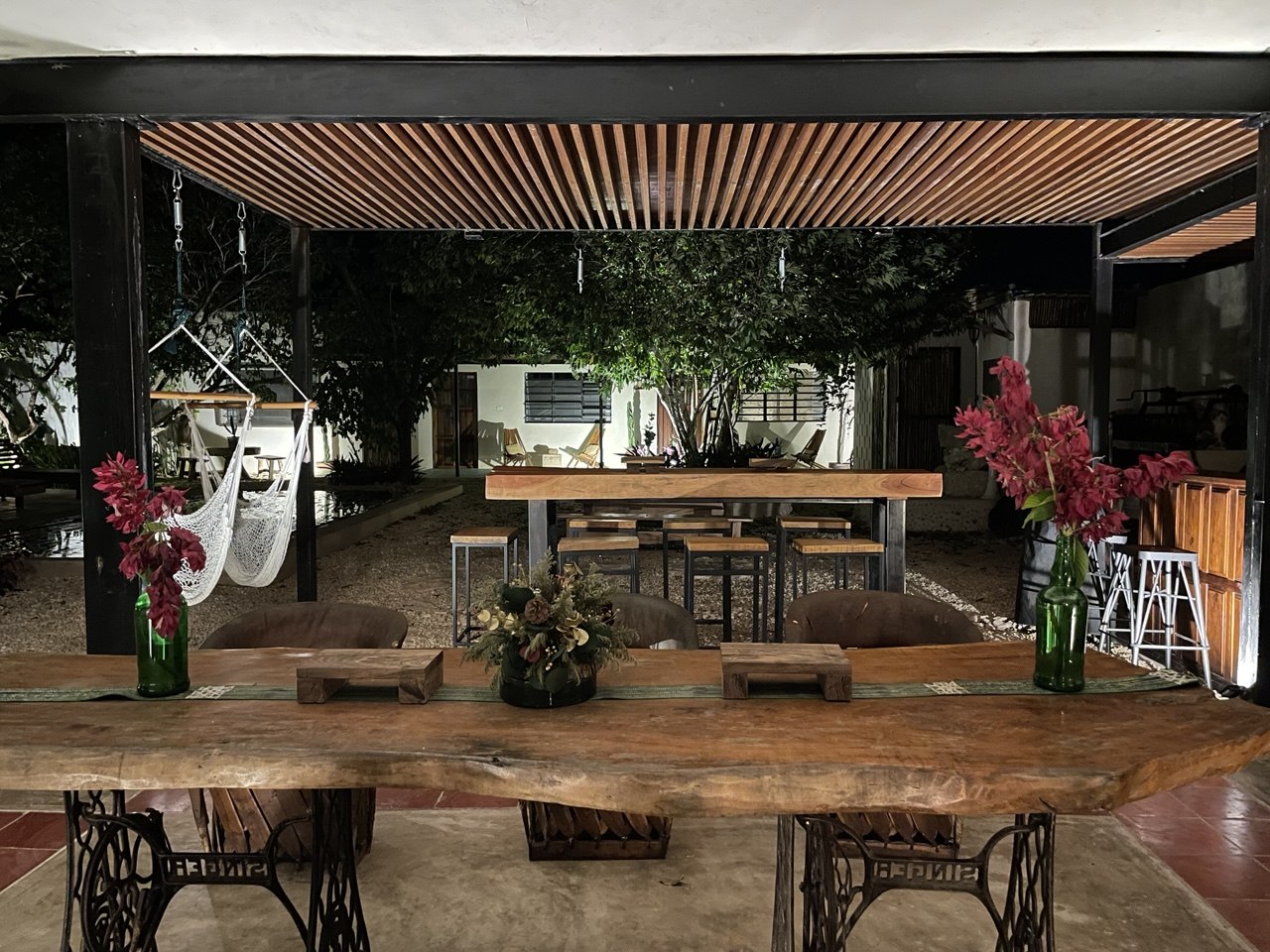 Patio exterior con mesa de madera rústica, hamacas y vegetación iluminada por la noche.