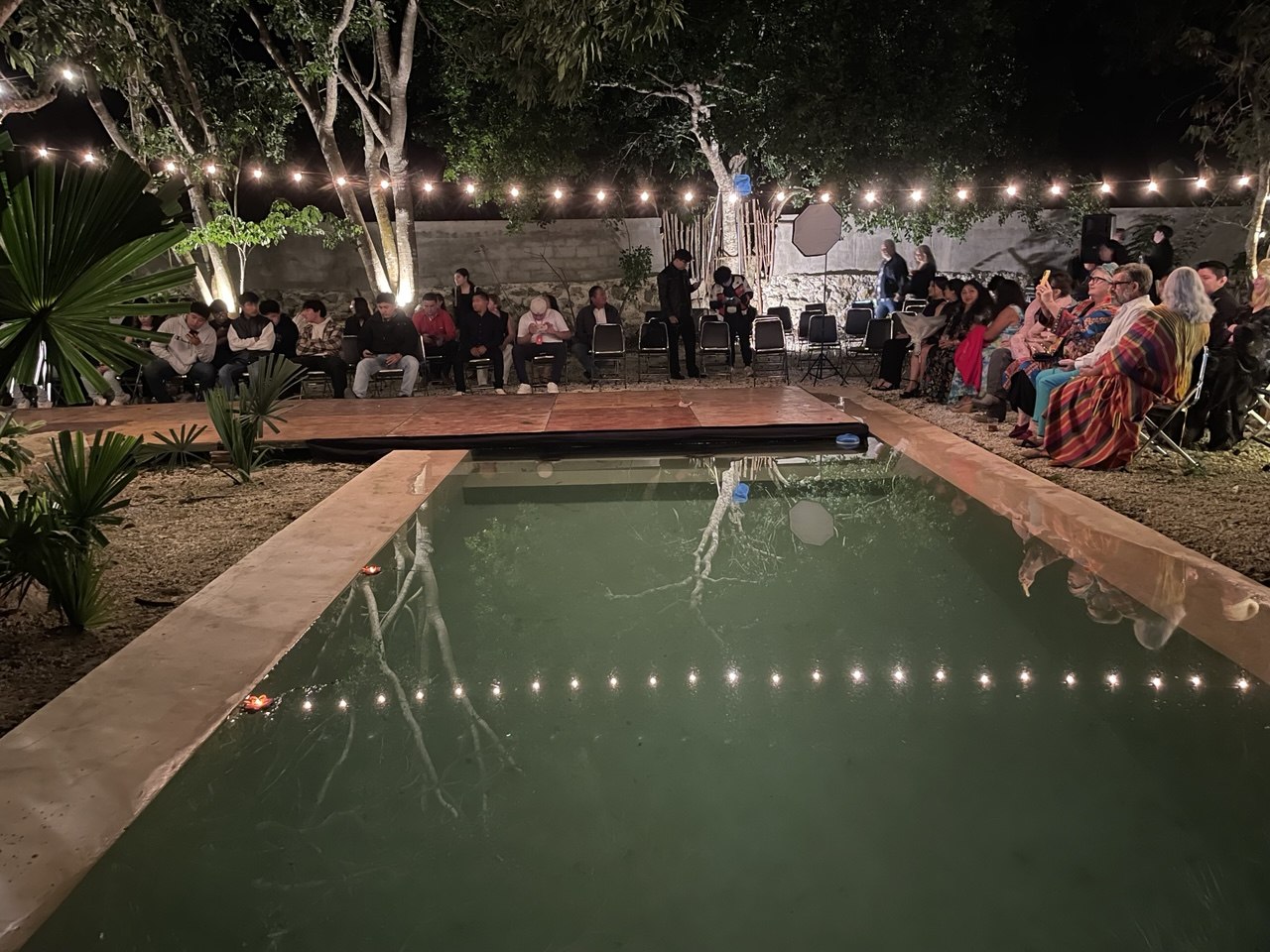 Alberca iluminada de noche con personas sentadas alrededor en un evento al aire libre.
