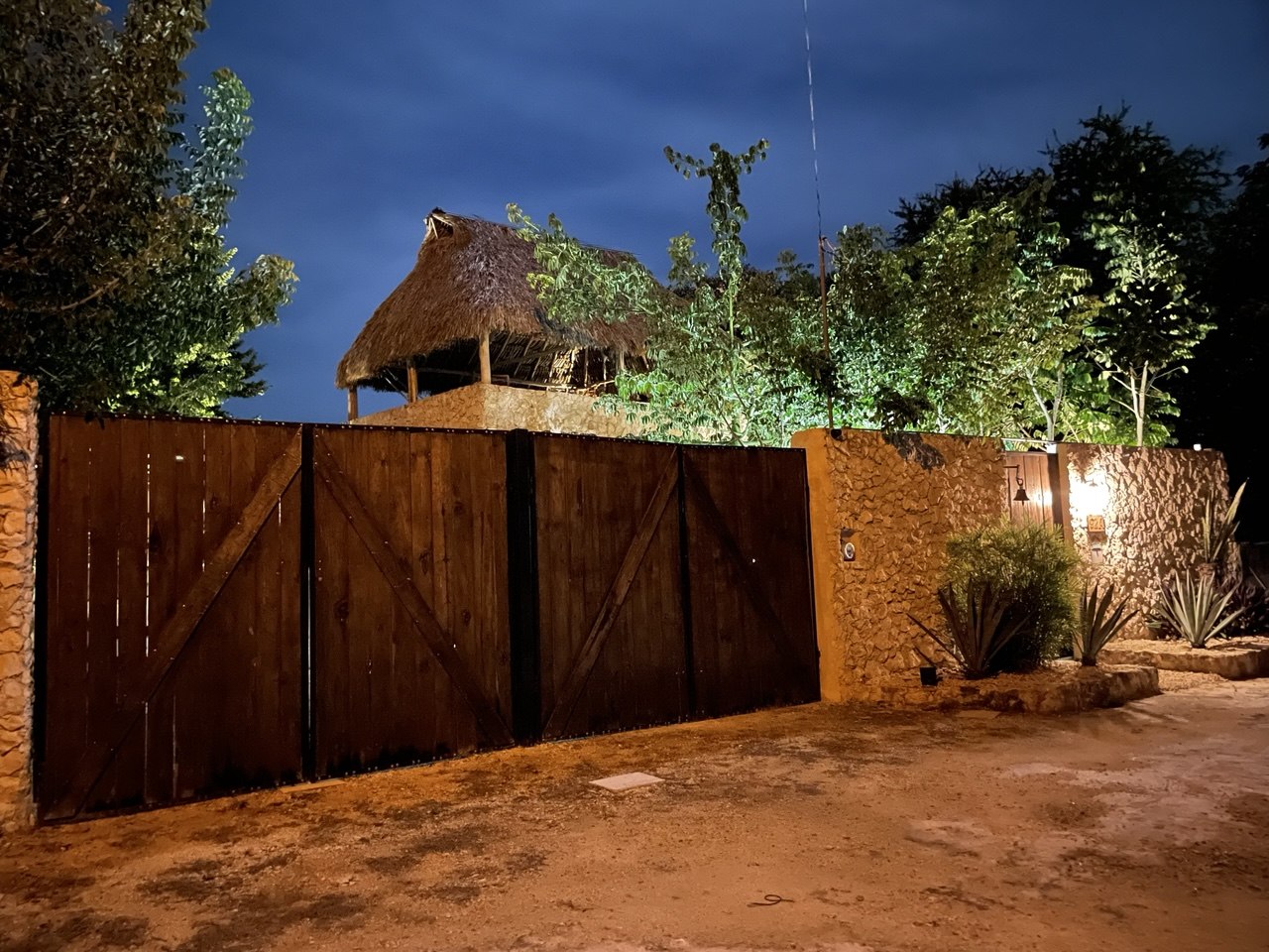 Vista nocturna de una propiedad con portón de madera, muro de piedra y techo de palapa.