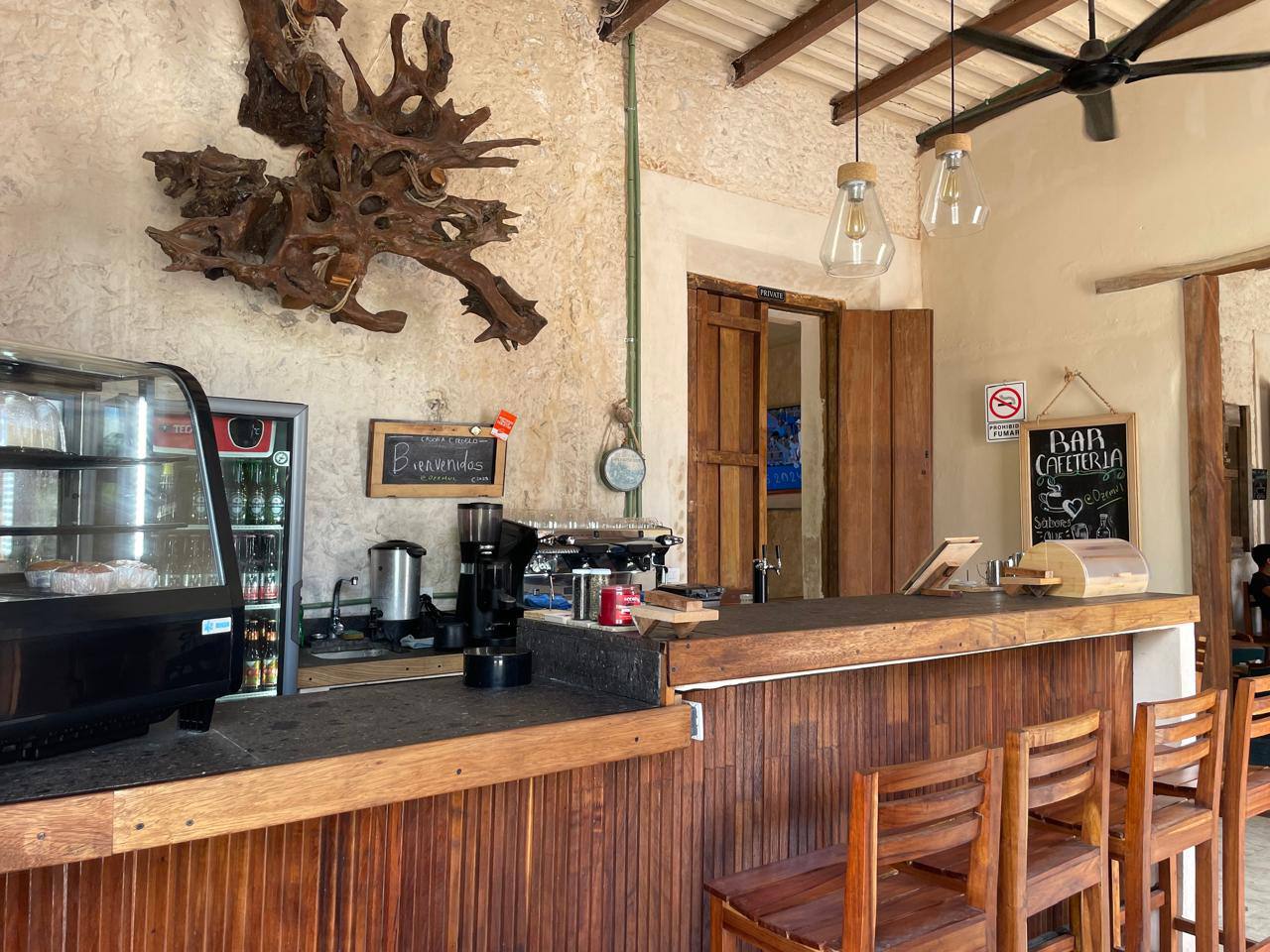 Barra de madera con taburetes, cafetera, nevera y decoración rústica en un local.