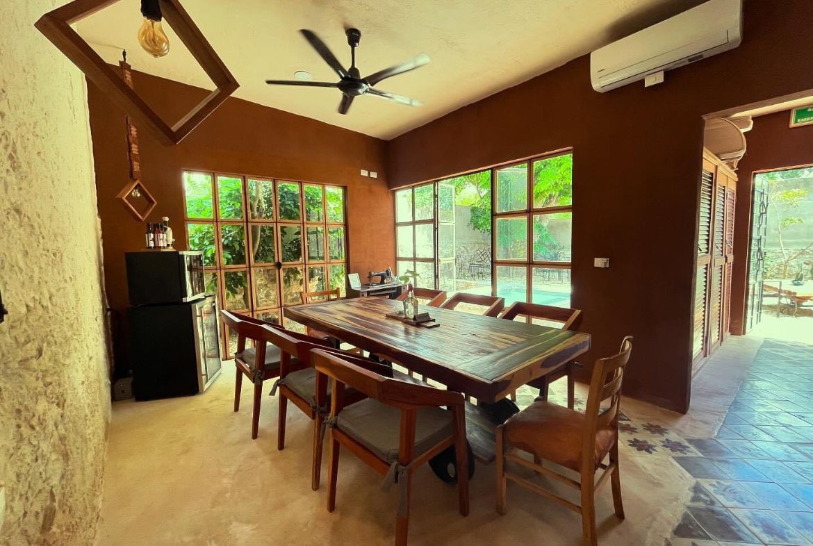Comedor con mesa de madera rústica, sillas, ventanas grandes y paredes marrones.