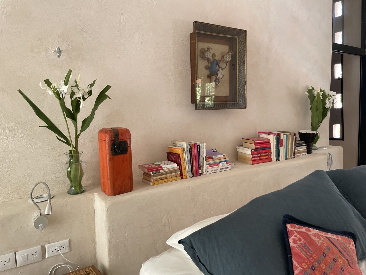 Cabecera de cama con libros, flores en jarrones y un cuadro decorativo en la pared.