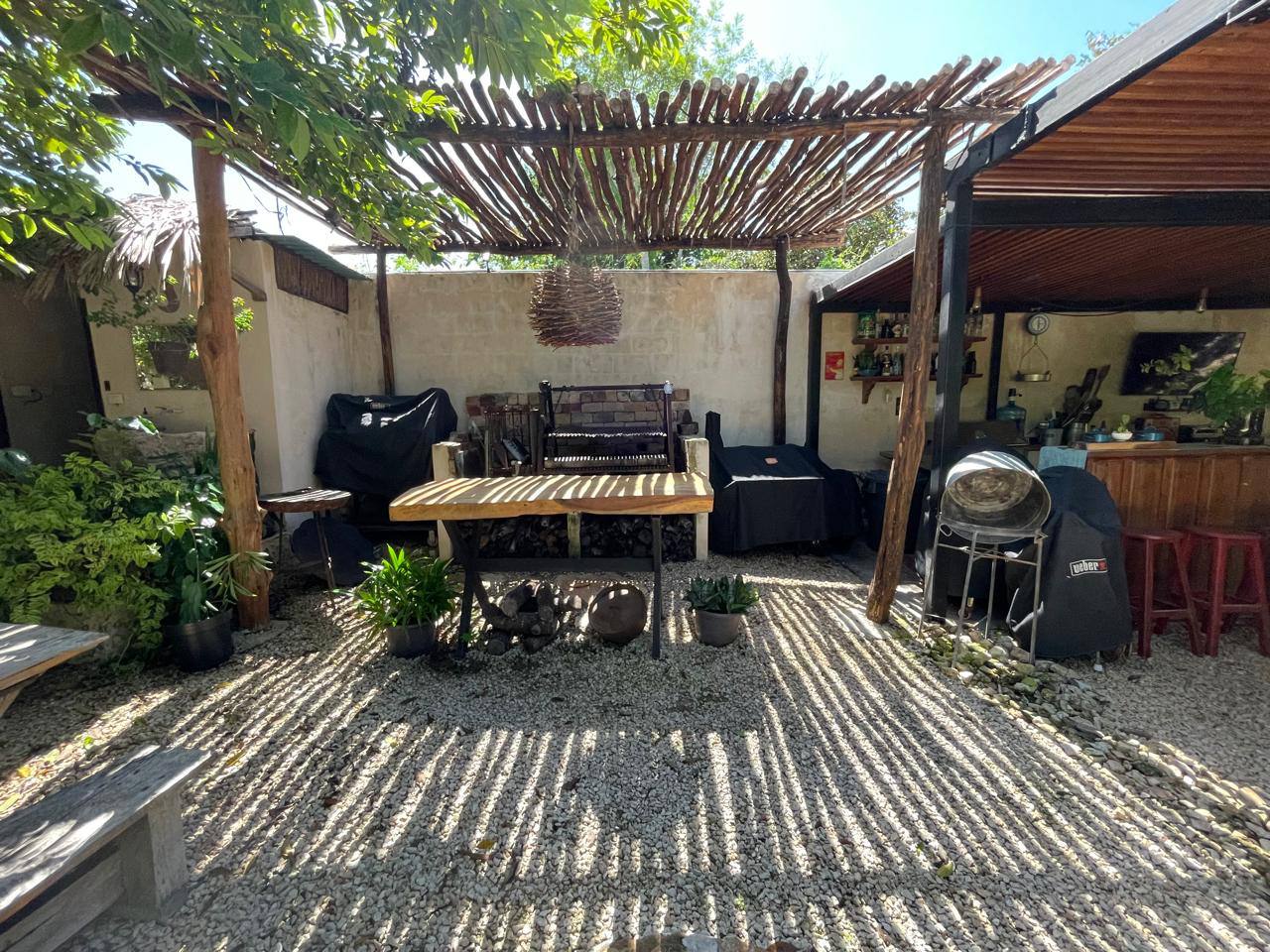 Patio exterior con techo de pérgola, parrillas cubiertas, mesa de madera y suelo de grava.