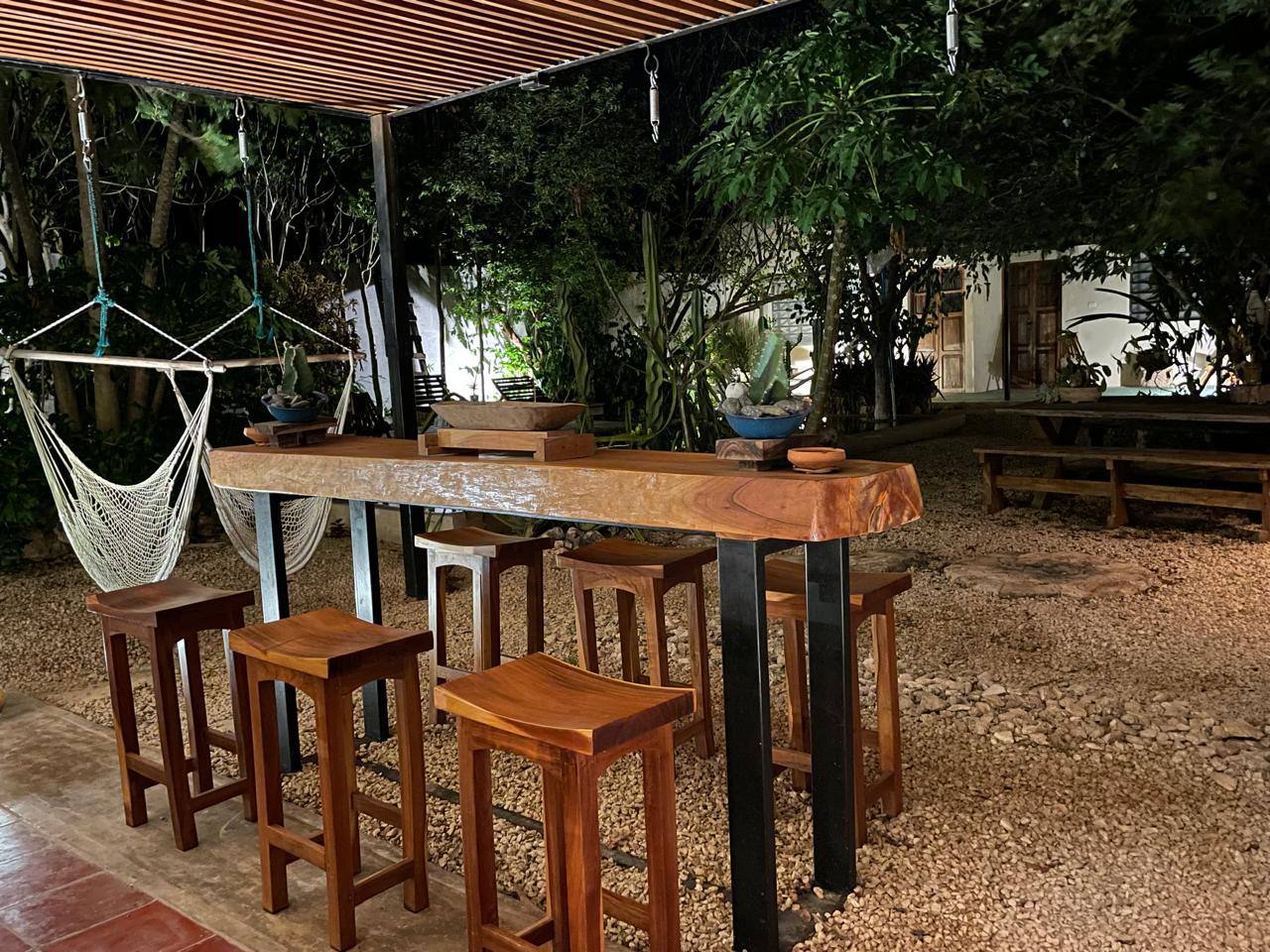 Patio exterior con barra de madera, taburetes, hamaca y área de jardín con árboles.